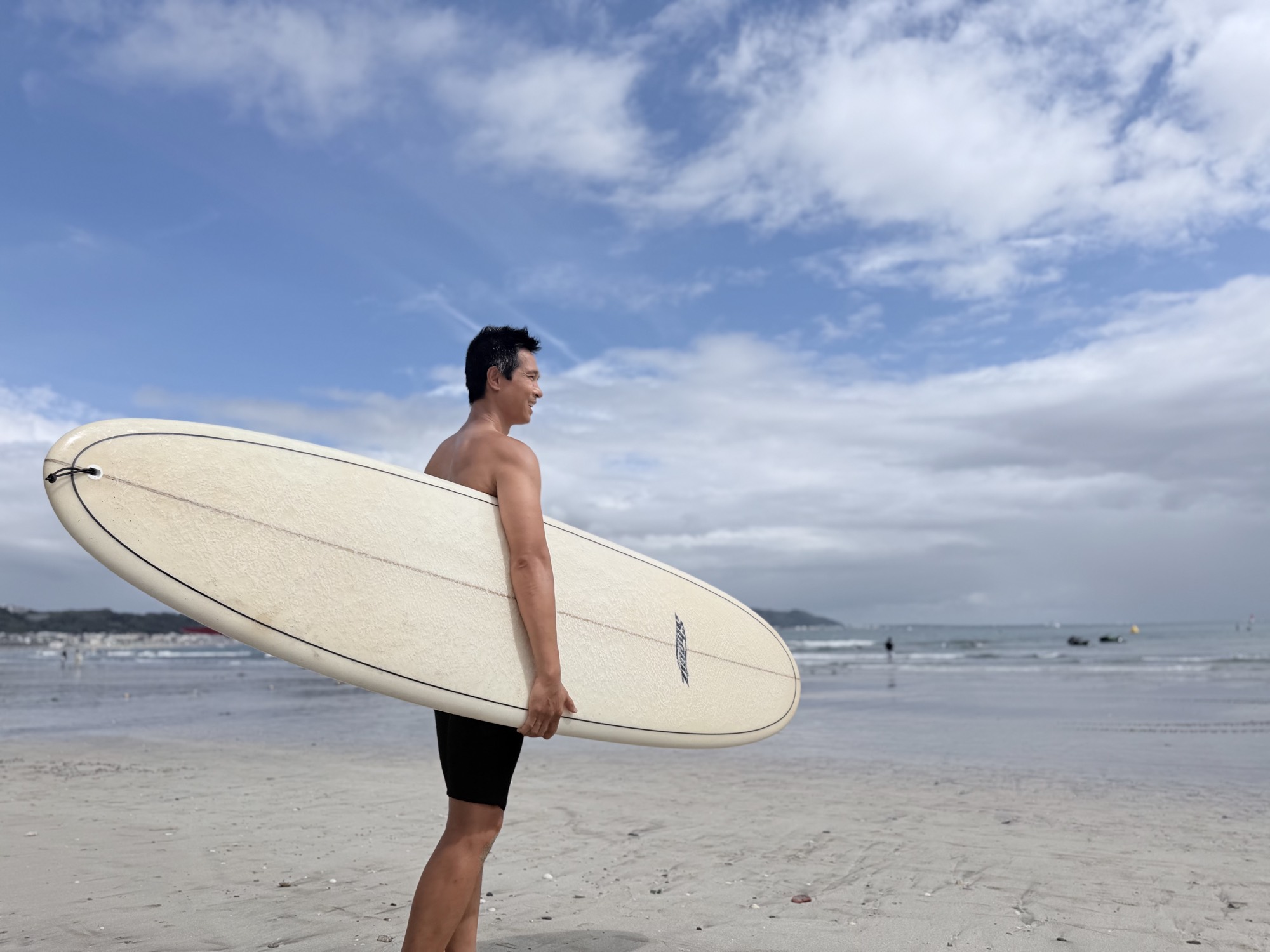 The host carrying a longboard on a Kamakura beach under a wide blue sky